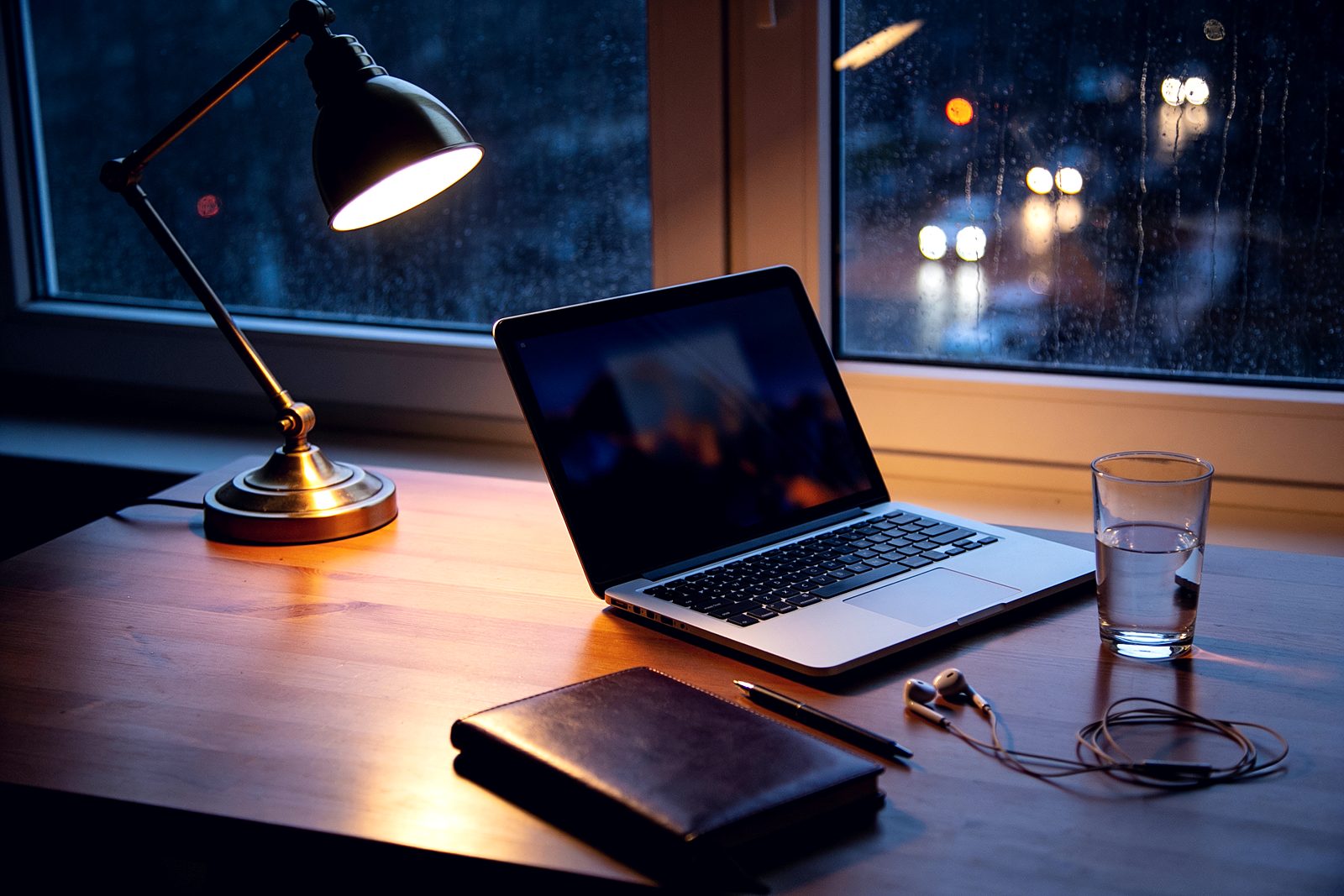A single laptop on a desk at night, lit by one lamp, rain on the window behind