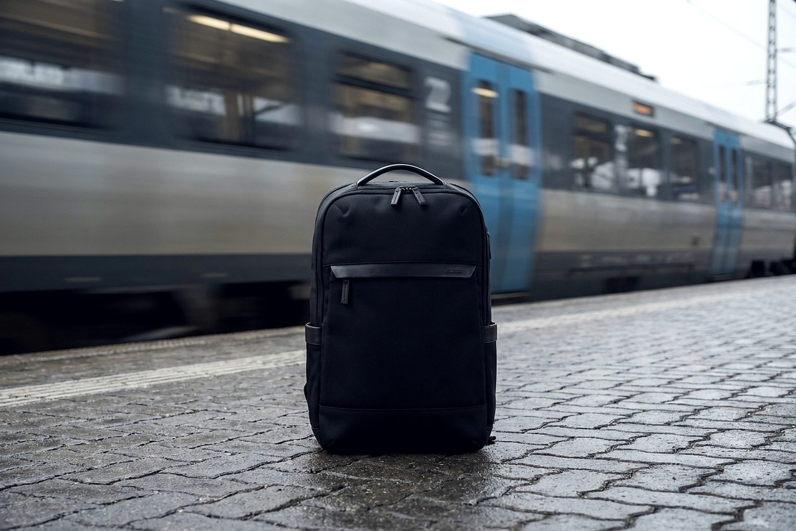 A structured black backpack standing on a railway platform, a train passing in motion blur behind