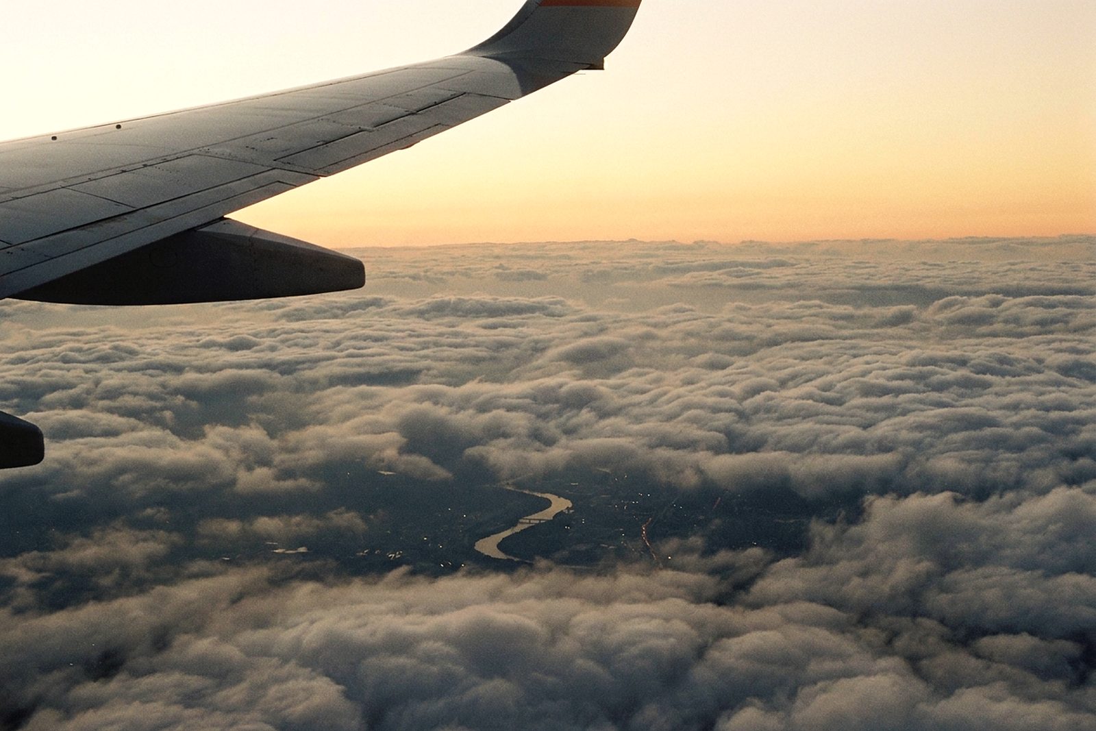 An aircraft wing above layered clouds at sunset, a river city visible far below