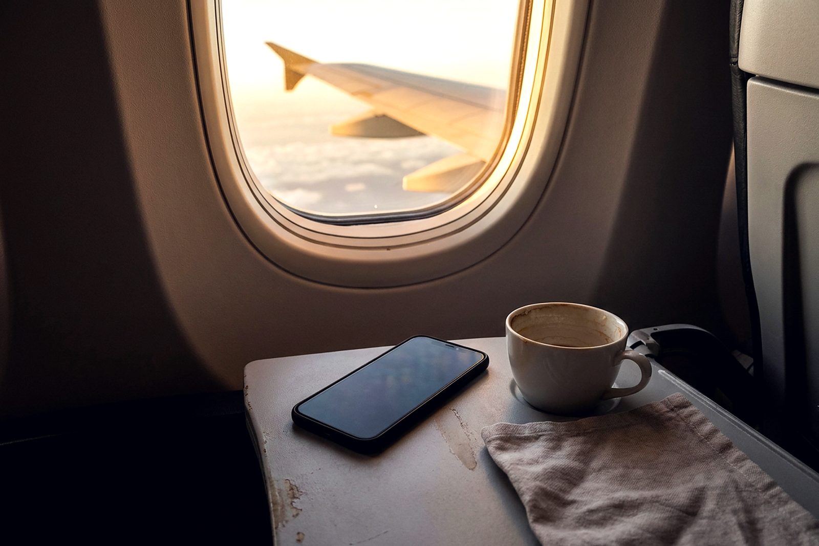A smartphone face-down on an aircraft tray table beside a window, the wing golden outside