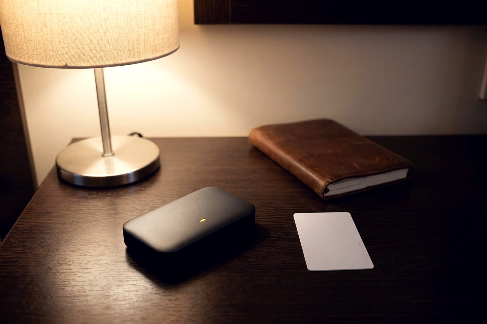 A pocket travel router with a single glowing light on a dark hotel desk beside a room keycard